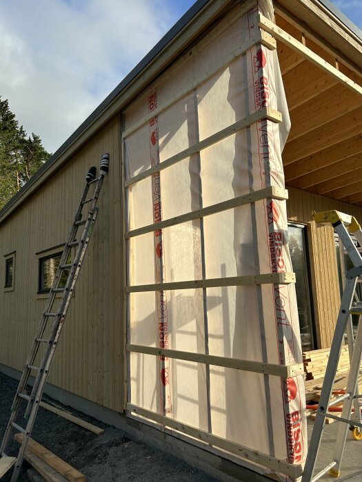 Trähus under konstruktion med isolering, vindskydd, stege och byggnadsmaterial synligt. Soligt väder.