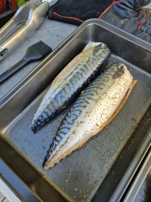 Två kryddade fiskar på en bakplåt bredvid grilltänger och pensel. Utomhus matlagning.