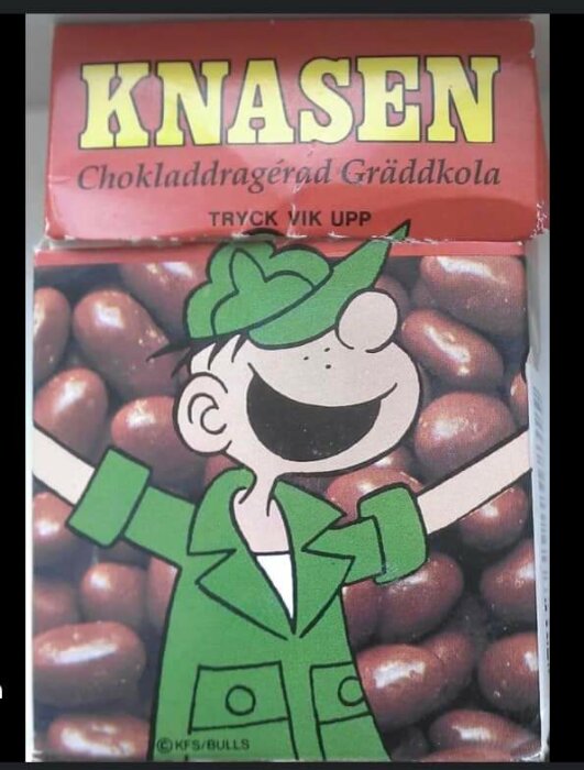 En bild på en godisförpackning med tecknad figur, märkt "KNASEN", chokladdrageér, gräddkola.