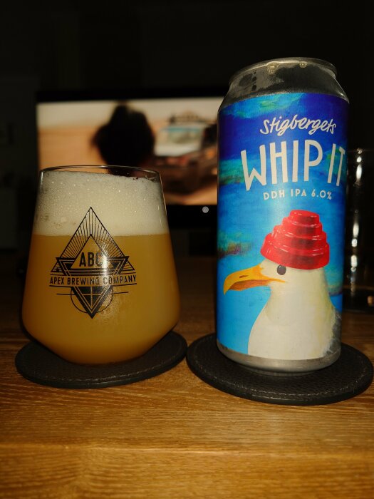 Ölglas och burk på träbord framför suddig TV-skärm. Apex Brewing-logga. Stigbergets bryggeri. "Whip It" DDH IPA.