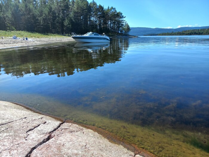 Klart vatten, motorbåt, strand, skog, berg, solig dag.