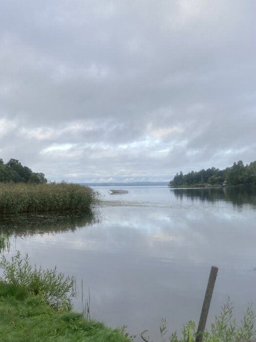 Ett stilla vattenlandskap med disiga himlar, speglingar, en enslig båt och vass vid strandkanten.