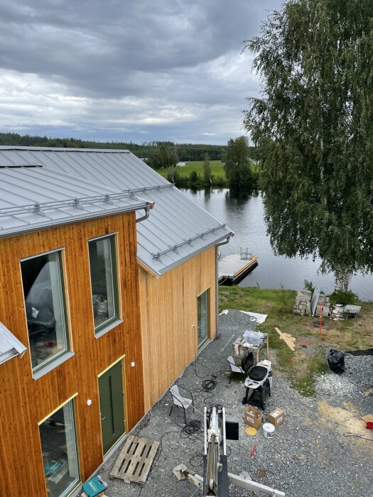 Trähus vid vatten med båt, molnig himmel, byggmaterial, pågående konstruktionsarbete, gröna dörrar, grått tak.