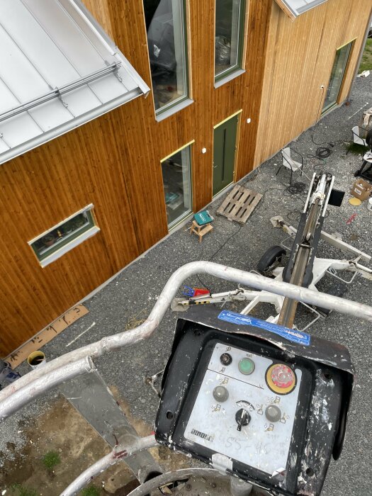 Utsikt från en lift vid en träbeklädd byggnad, oordning på marken, byggmaterial och skräp syns.