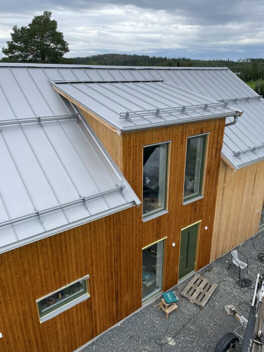 Trähus med silverfärgat plåttak, skog i bakgrunden, molnig himmel, några fönster, säkerhetsräcke på taket.