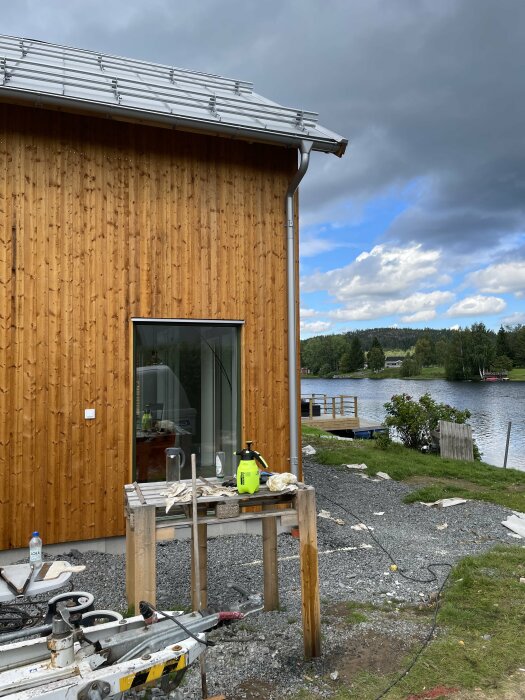 Trähustillbyggnad under arbete, verktyg och material framför, mörka moln över vattnet i bakgrunden.