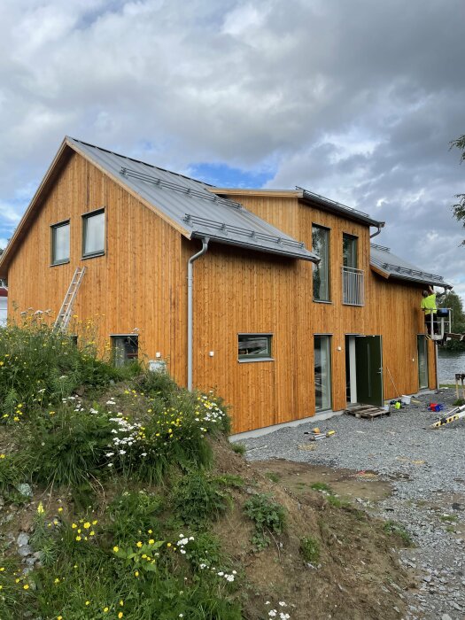 Nybyggt trähus med stående panel, metalltak, stegar, under arbete, delvis trädgårdsanläggning, molnig himmel.