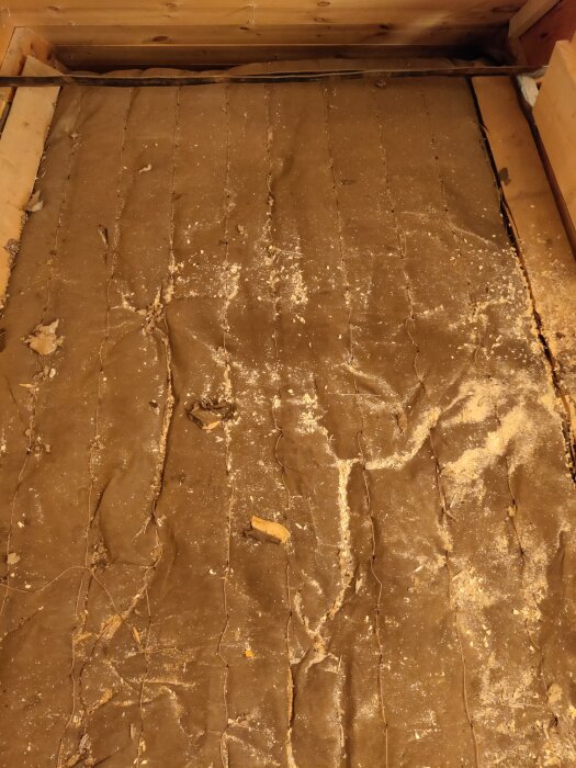 Ett smutsigt golvtäcke av textil med sprickor och skräp; möjligen ett källar- eller vindsgolv.