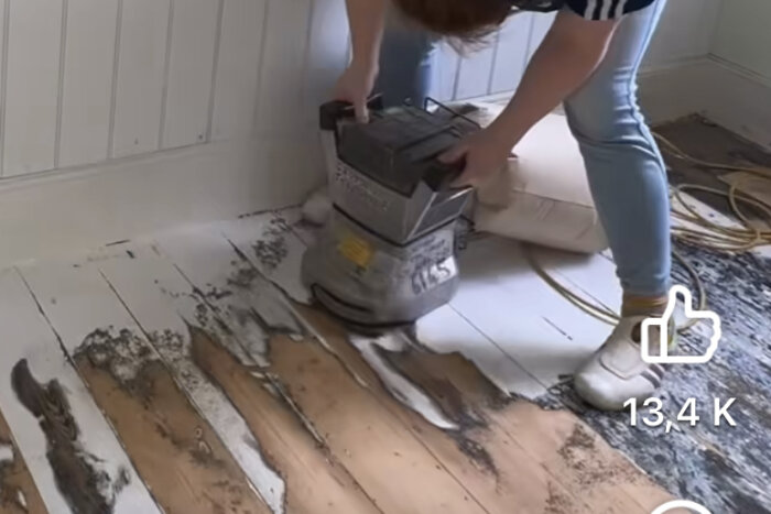 Person slipar trägolv, bär på slipmaskin, gammalt skadat golv syns, renoveringsarbete pågår.