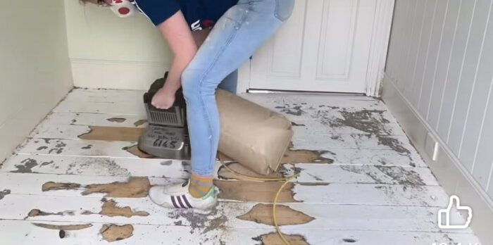 Person böjer sig ner i rum med skadat trägolv, troligen renoveringsarbete pågår.