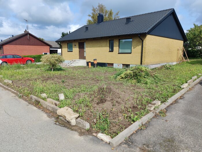 Gult enplanshus med svart tak, oanlagd trädgård, röd bil, molnig himmel, bostadsområde.