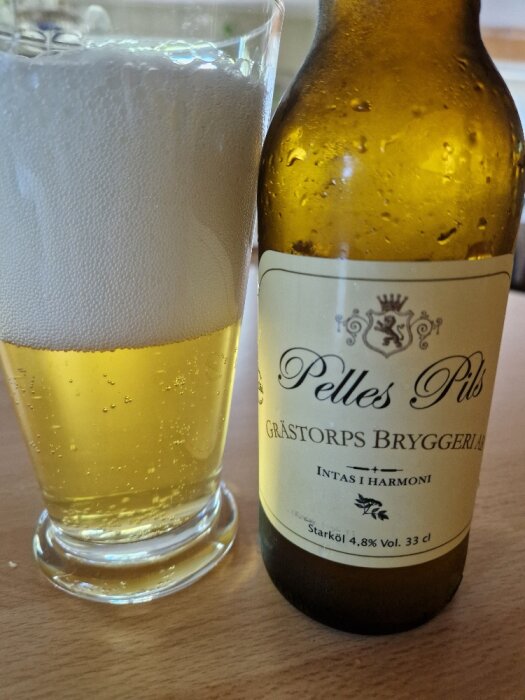 Ett glas med öl och en delvis synlig ölflaska märkt "Pelles Pils, Grästorps Bryggeri".