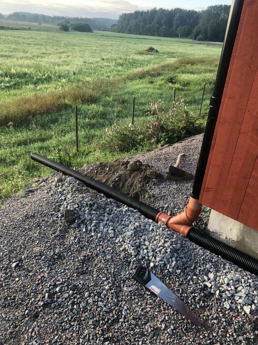 Ett grönt fält vid gryning, avloppsrör nära en röd byggnad, såg och sten på marken.