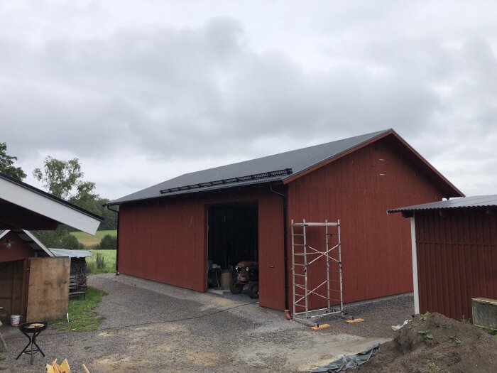 Röd lada med öppna dörrar, byggnadsställning bredvid, molnig himmel, grusgång, byggmaterial på marken.