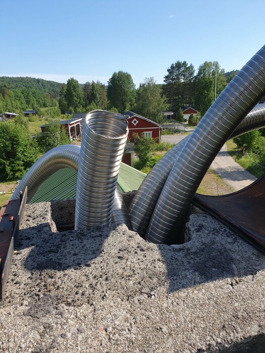 Flexibla ventilationsrör sticker ut från tak, omgiven av grönska och röda hus. Solljus och klarblå himmel.