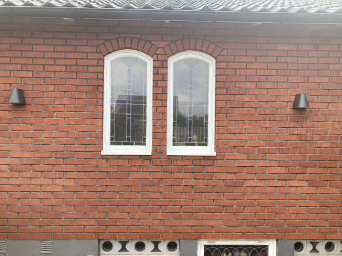 Två vita bågformade fönster i tegelvägg, utomhuslampor, del av grå grund.