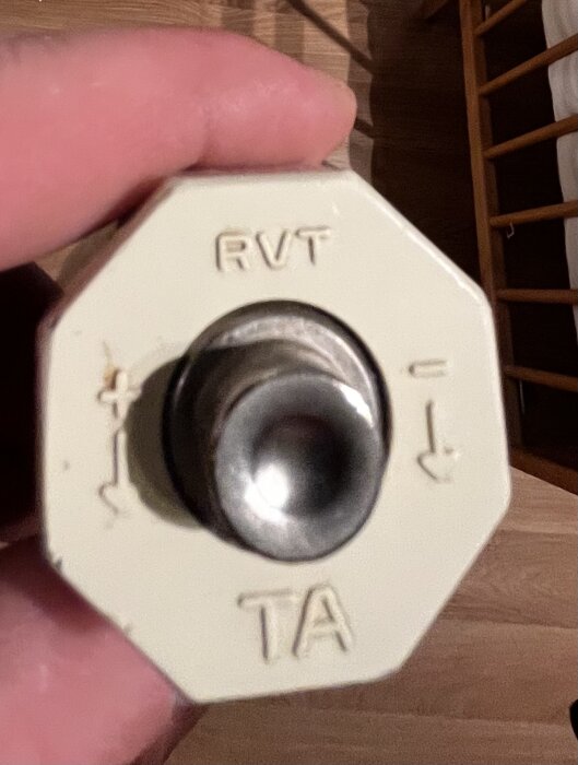 Åttkantig vit enhet hållen i hand, text "RVT" och "TA", pilmarkeringar, justeringsmekanism eller sensor i mitten.