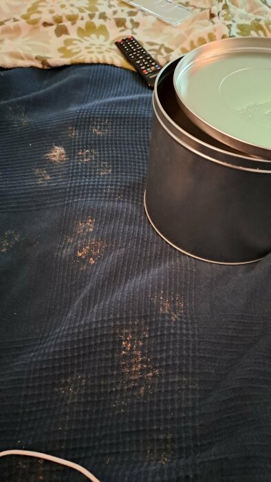 Svart behållare, fjärrkontroll, kuddar, blått tyg med hår och smutsfläckar.