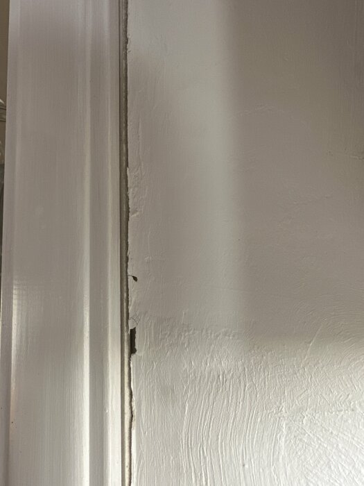 Vit målad dörrkarm och vägg med synligt slitage och reflekterande ljusstrimma.
