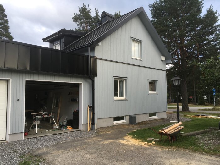 Tvåvåningshus, grå fasad, öppet garage, verktyg, byggnadsmaterial, träd, gräsplätt, övergivet barnvagn, molnig himmel.