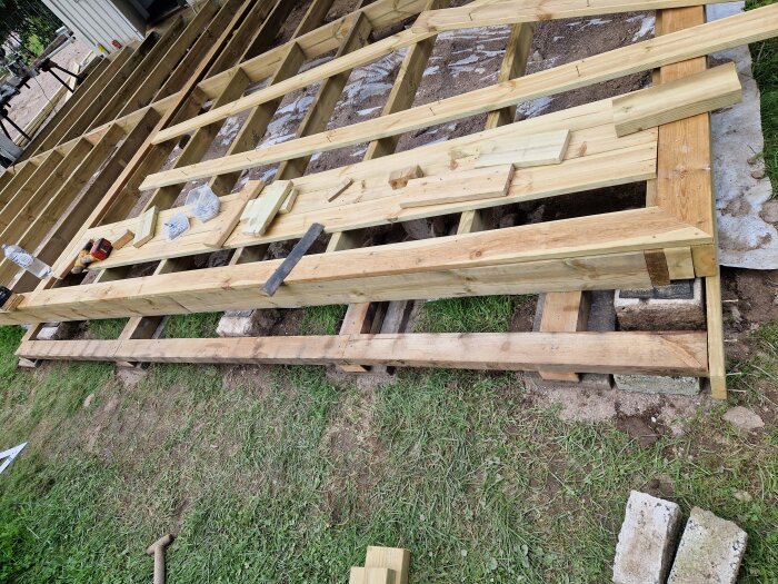 Pågående konstruktion av trästruktur, material och verktyg på marken, oklart projektstadium, utomhusbygge.