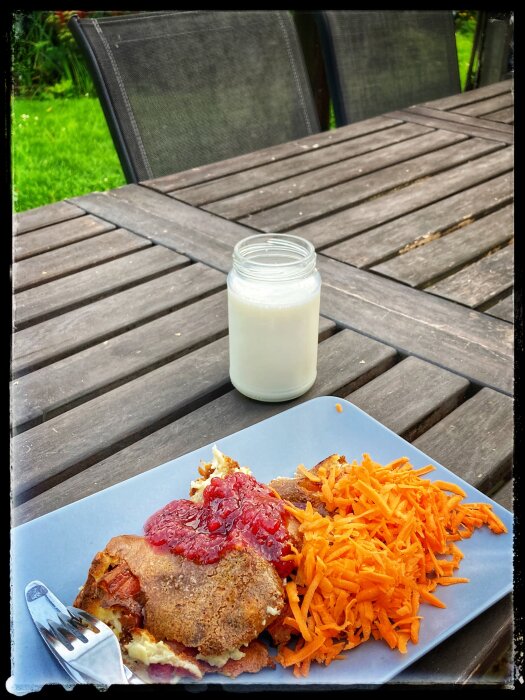 Måltid bestående av pannkaka, sylt och riven morot på blå tallrik, glas med mjölk, träbord utomhus.