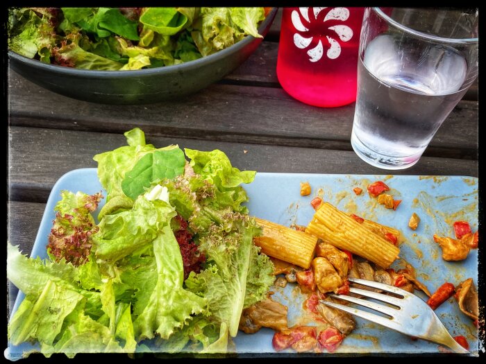 Nästan färdigätten måltid bestående av pasta och sallad, vattenglas, träbord, utomhuslunch eller middag.