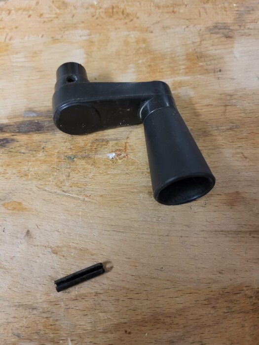 Svart plastdel och pinne på träyta, del kanske till cykel eller maskin, verktygs- eller utrustningskomponent, slitage synligt.