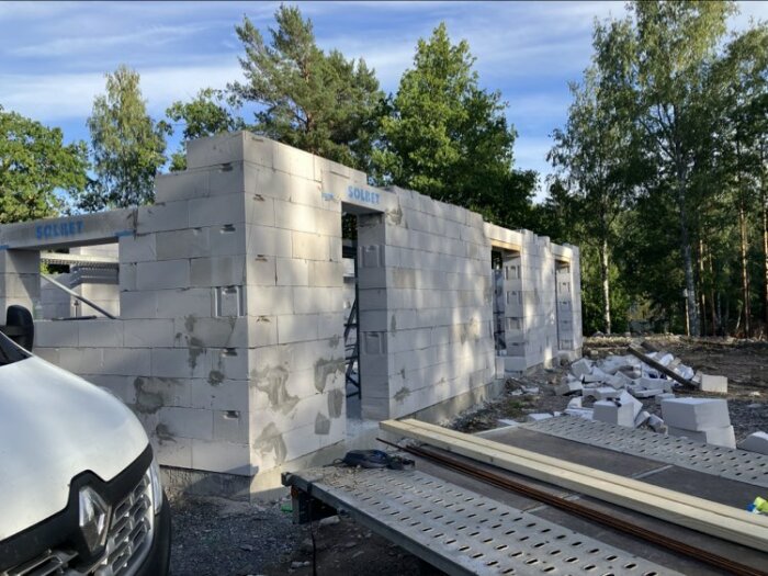 Byggarbetsplats med ofärdiga väggar, grå betongblock och byggmaterial, skog i bakgrunden, del av fordon framför.