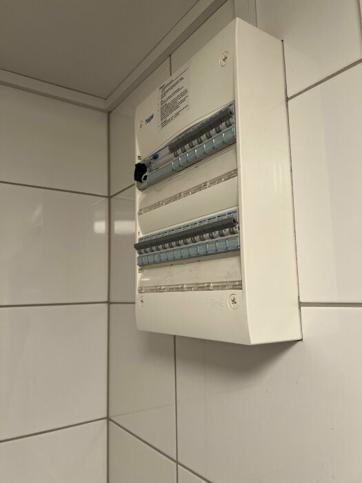 Vit elcentral med säkringar på kakelvägg, etikett, säkringsomkopplare i olika positioner.
