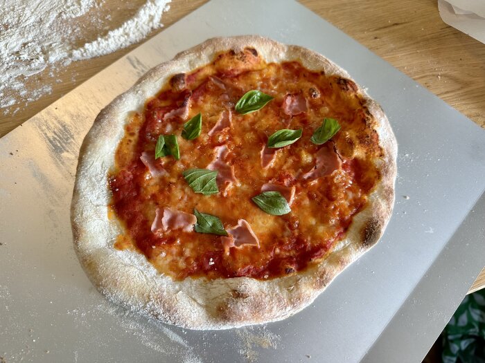 Nybakad pizza med tomatsås, ost, skinka, basilika på träbord med mjöl.