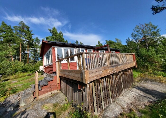 Röd stuga med veranda på klippa i skogsmiljö, blå himmel, solig dag.