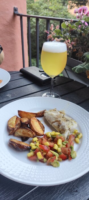 Middag på balkong: grillad fisk, klyftpotatis, tomatsalsa, ölglas. Grönt, blommor i bakgrunden. Sommartid, avkoppling.