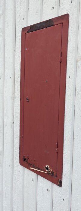 Röd dörr eller lucka med rost och slitage, på vit trävägg, stängd och låst, vertikal komposition.
