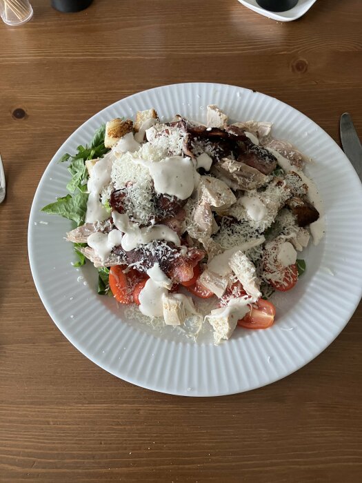 Caesarsallad med kyckling, bacon, krutonger, parmesan, romansallad, dressing på tallrik, träbord.