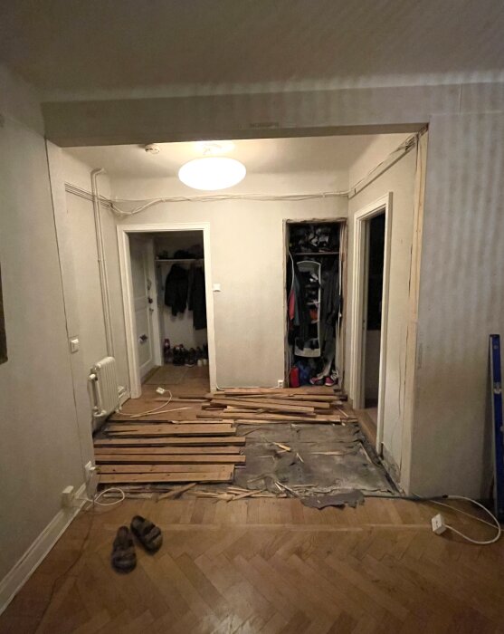 Renovering pågår, trägolv tas upp, hög med plankor, sladdar på golvet, garderob i bakgrund, inomhusbelysning.