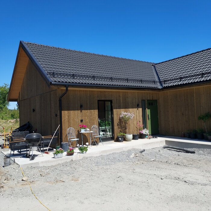 En trähusfasad med terrass, grill, stolar och krukväxter under en klarblå himmel.