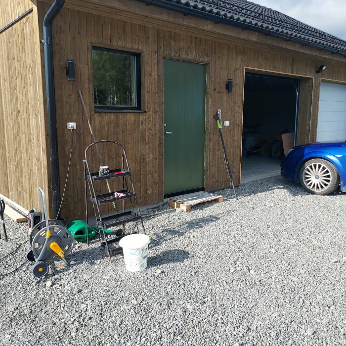 Träfasad byggnad, dörr, garage öppet, bil, stegar, utomhus elektrisk utrustning, grusuppfart, soligt väder.