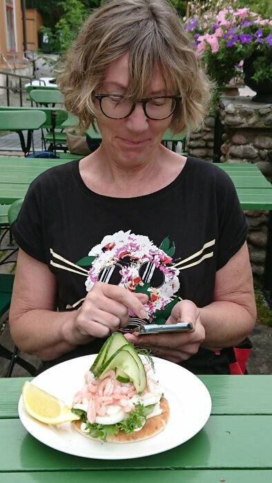 Kvinna med glasögon vid grönt bord ser på telefon, framför räksmörgås med citron och gurka.