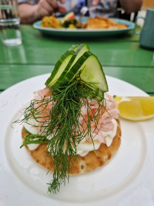 Räksmörgås med dill, gurka, majonnäs på en vit tallrik, citronklyfta, uteservering.