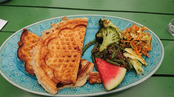 Färgrik tallrik med våfflor, grönsaker, vattenmelon, morotsallad på grönt bord.