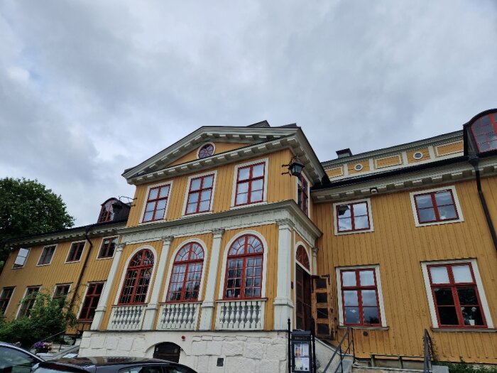 Historisk träbyggnad med stenvåning, röda fönsterluckor, parkerade bilar, mulet himmel.