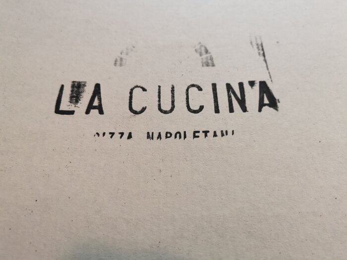 Svart text "LA CUCINA" på en suddig, fläckig vit bakgrund, med oskarpa små texter nedanför.