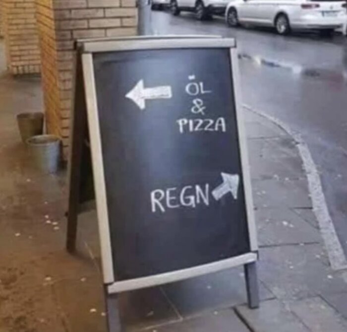 Svart skylt med text "ÖL & PIZZA" och "REGN", pilar pekar, vått trottoar, vägg, bilar.