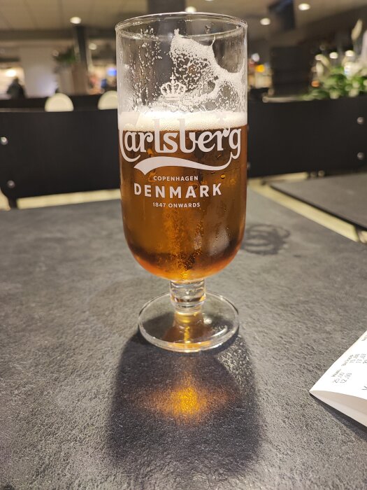 Ett halvdrucket glas Carlsberg öl på ett bord i en inomhusmiljö.