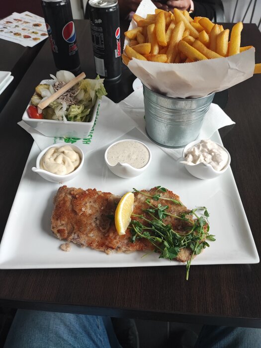 Måltid med schnitzel, pommes frites i hink, sallad, såser, och läsk, serverat på ett bord.