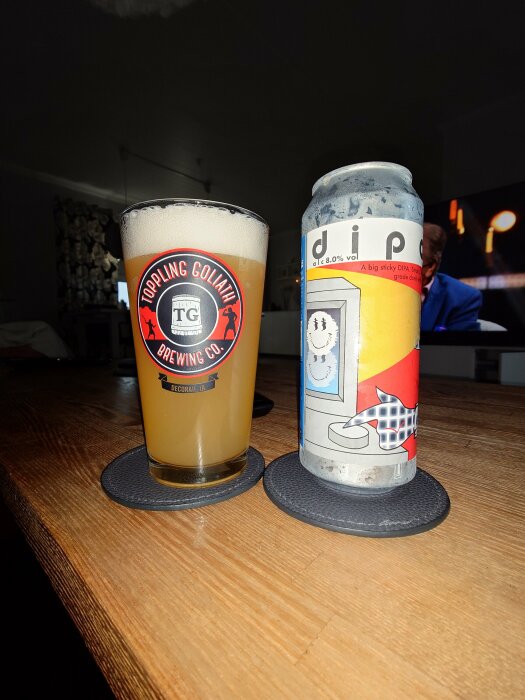 Ett glas öl och en ölburk på bord, Toppling Goliath Brewing Company, inomhus, bordsunderlägg, svagt ljus.