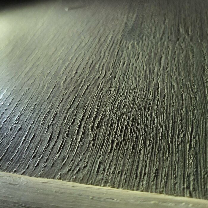 Närbild på en texturerad yta som liknar mörkt trä eller träimitation med tydliga linjer och kornighet.
