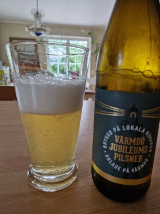 Ölglas med skum, flaska etikett "Värmdö Jubileums Pilsner", hemmiljö, fönster, ljust, inomhus.