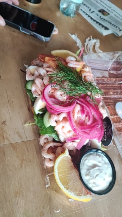 Skaldjursrätt med räkor, citron, dill och picklad rödlök på en skärbräda; person fotograferar maten.